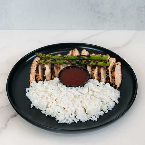 The Arnold 2022 Bowl features sliced grilled chicken, white rice, grilled asparagus, and a dark dipping sauce in a sleek black plate—ideal for a healthy lunch or easy chicken and rice meal prep.