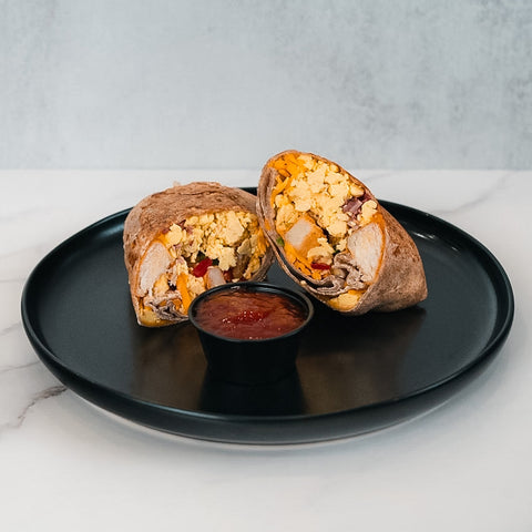 The Breakfast Burrito, filled with scrambled eggs, turkey bacon, and vegetables, is cut in half and served on a black plate with a small container of red sauce in the center.