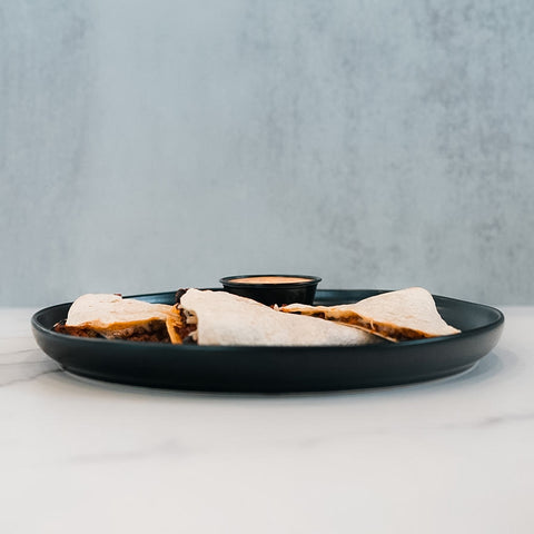 A black plate holds four Vegan Chorizo Quesadilla wedges and a small cup of sauce, perfect for dinner, set on a white surface with a light gray background.