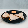 Three slices of Vegan Chorizo Quesadilla, filled with hearty beans, are served on a black plate with orange dipping sauce, set against a light gray wall and white marble surface—ideal for a tasty dinner or healthy lunch.