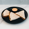 Three Vegan Chorizo Quesadilla wedges, filled with plant-based chorizo and beans, are arranged on a black plate with orange sauce in the center—ideal for a healthy lunch or delicious dinner—set on white marble.