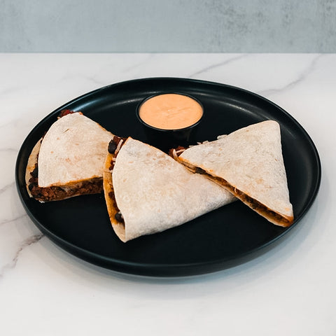 Three Vegan Chorizo Quesadilla wedges, filled with plant-based chorizo and beans, are arranged on a black plate with orange sauce in the center—ideal for a healthy lunch or delicious dinner—set on white marble.
