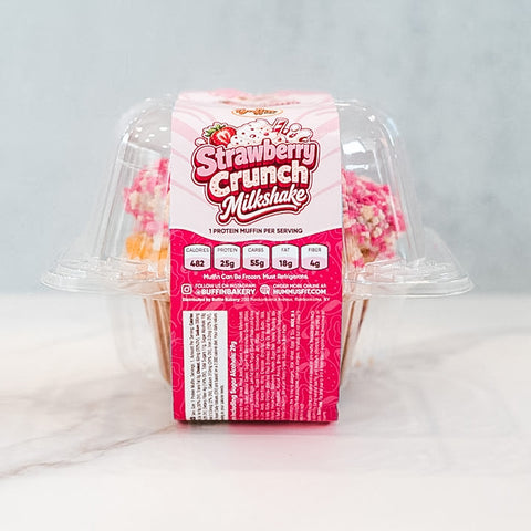 A clear plastic container holds Strawberry Crunch Milkshake - Exclusive- 20 protein muffins from Buffin Bakery, featuring a pink label with nutrition facts and branding, displayed on a white surface against a light background.