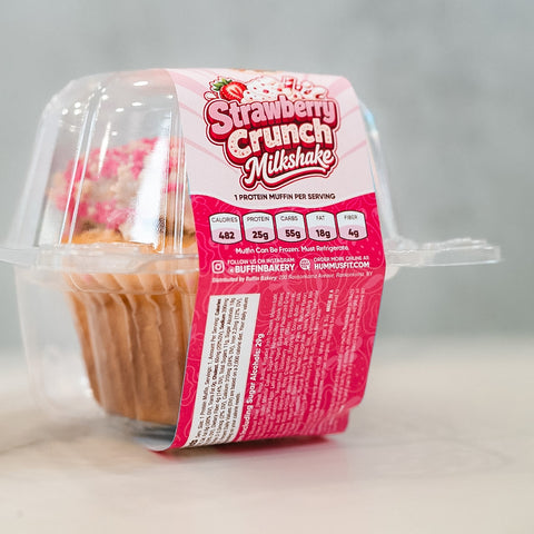 A clear container holds a high protein Strawberry Crunch Milkshake Buffin Muffin, featuring a pink label with the product name. The label displays nutrition facts and branding, while the background appears light and blurred.
