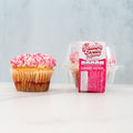 A single cupcake with pink and white sprinkles sits next to a clear plastic container labeled Strawberry Crunch Milkshake Buffin Muffin, which holds a high protein muffin with a delicious strawberry milkshake flavor.