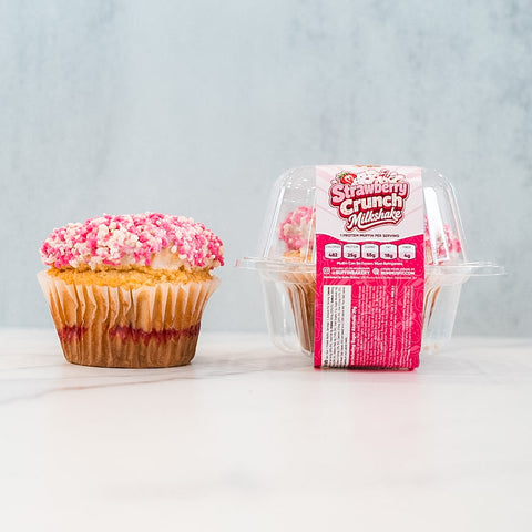A single cupcake with pink and white sprinkles sits next to a clear plastic container labeled Strawberry Crunch Milkshake Buffin Muffin, which holds a high protein muffin with a delicious strawberry milkshake flavor.