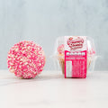 A Strawberry Crunch Milkshake Buffin Muffin sits next to its clear plastic packaging, which is labeled Strawberry Crunch Milkshake Buffin Muffin. Featuring a strawberry milkshake flavor, this buffin muffin is coated in a pink and white crunchy layer.