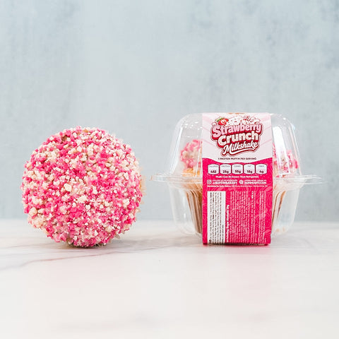 A Strawberry Crunch Milkshake Buffin Muffin sits next to its clear plastic packaging, which is labeled Strawberry Crunch Milkshake Buffin Muffin. Featuring a strawberry milkshake flavor, this buffin muffin is coated in a pink and white crunchy layer.