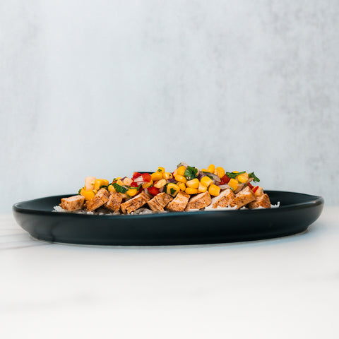 The Southwest Chicken Bowl features diced grilled chicken topped with corn, chopped tomatoes, and fresh herbs, all served in a black bowl on a white surface against a light gray background.