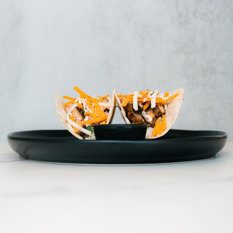 Baja Chicken Tacos feature grilled chicken, shredded cheddar, and mozzarella cheese in low-carb flour tortillas, served on a black plate with dipping sauce at the center, set against a light gray background.
