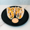 Baja Chicken Tacos: Two grilled chicken soft tacos with cheddar, white cheese, and herbs on low-carb flour tortillas. Served on a black plate with creamy dipping sauce. Ideal for high-protein meal prep.