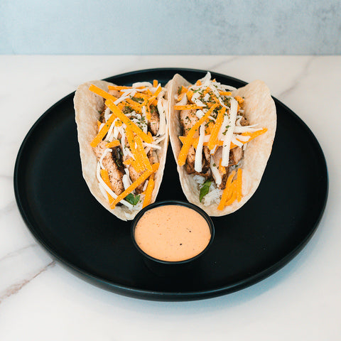Baja Chicken Tacos: Two grilled chicken soft tacos with cheddar, white cheese, and herbs on low-carb flour tortillas. Served on a black plate with creamy dipping sauce. Ideal for high-protein meal prep.