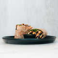 The Grilled Chicken Pesto Wrap, halved and filled with greens and grilled chicken, is drizzled with sauce and served on a black plate with a small dipping cup—ideal for meal prep.