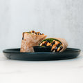 A Grilled Chicken Pesto Wrap, sliced to show fresh greens and grilled chicken, is served on a black plate with a side of dark dipping sauce.