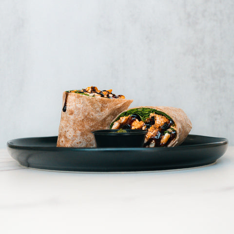 A Grilled Chicken Pesto Wrap, sliced to show fresh greens and grilled chicken, is served on a black plate with a side of dark dipping sauce.