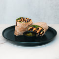 The Grilled Chicken Pesto Wrap features grilled chicken, fresh spinach, and a tasty pesto sauce, served halved on a black plate with a side of green pesto dip—ideal for convenient meal prep.