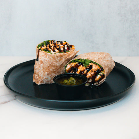 The Grilled Chicken Pesto Wrap, packed with protein and fresh greens, is cut in half and served with a side of green sauce—an ideal meal prep option for busy days.