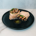 The Grilled Chicken Pesto Wrap features healthy grilled chicken, greens, beans, and veggies with pesto and balsamic glaze, sliced in half and served meal prep style on a black plate with a side of green sauce.