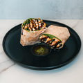 The Grilled Chicken Pesto Wrap, packed with protein, greens, and grilled chicken, is cut in half and drizzled with dark sauce on a black plate beside a small dish of green dipping sauce.