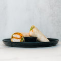 A black plate holds a Crispy Baked Chicken Wrap, halved and filled with lettuce and sauce, plus a small black container of dip in the center. Set on a light textured surface, it’s a high-protein lunch ideal for healthy meal prep.