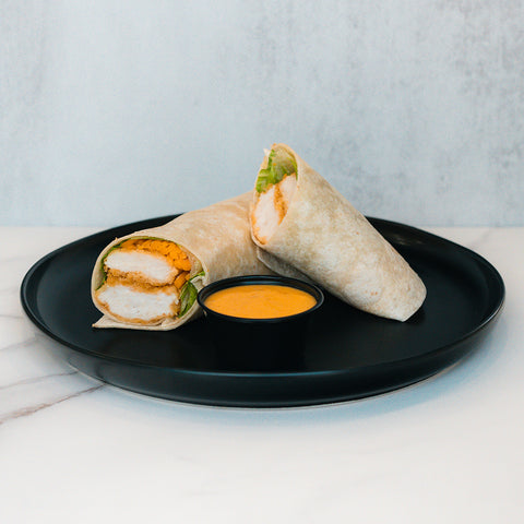 Two halves of the Crispy Baked Chicken Wrap, filled with lettuce and sauce, are served on a black plate with a small cup of dipping sauce—ideal for a high-protein lunch. The setup sits on a light-colored surface.