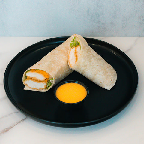 A black plate holds a Crispy Baked Chicken Wrap cut in half, showing breaded chicken, lettuce, and cheese. Served with a small cup of yellow dipping sauce, this wrap is ideal for healthy meal prep or a high-protein lunch.