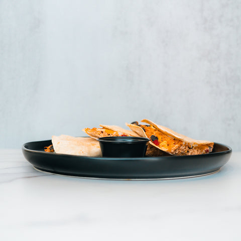 A black plate holds three slices of the Southwest Chicken Quesadilla, filled with cheese, Southwest-seasoned chicken, and vegetables—perfect for a healthy quesadilla meal prep. In the center is a small black cup of chipotle sauce. The background features a light, minimalist surface.