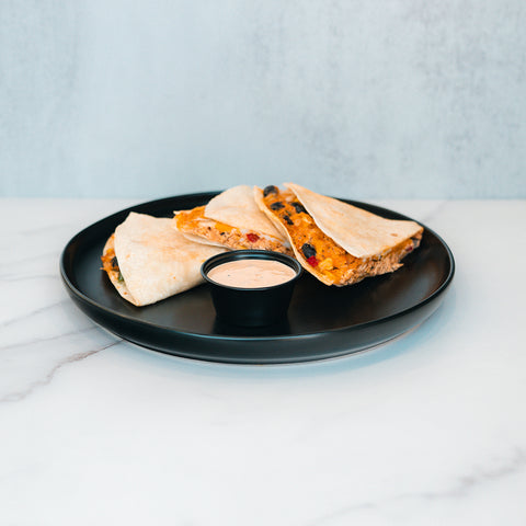 Three wedges of the Southwest Chicken Quesadilla, filled with melted cheese and vegetables, are arranged on a black plate alongside a small cup of chipotle sauce, set atop a white marble surface—perfect for healthy meal prep or enjoying as a flavorful spin on the classic quesadilla.
