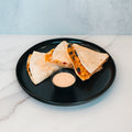 Three Southwest Chicken Quesadilla wedges with delicious filling are arranged on a round black plate and served with a small cup of creamy chipotle sauce on a white marble surface—making them an ideal healthy meal prep option for fans of the Southwest Chicken Quesadilla.