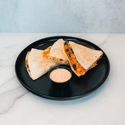 Three Southwest Chicken Quesadilla wedges with delicious filling are arranged on a round black plate and served with a small cup of creamy chipotle sauce on a white marble surface—making them an ideal healthy meal prep option for fans of the Southwest Chicken Quesadilla.