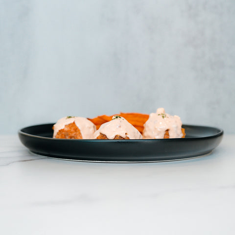 A black plate features Buffalo Chicken Meatballs—three breaded bites topped with a creamy sauce—served alongside a small portion of orange chips on a white marble surface with a light gray background, making them perfect for a healthy dinner or your next meal prep.