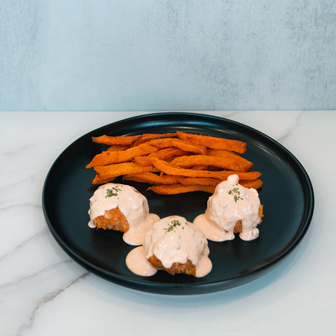 A black plate with three Buffalo Chicken Meatballs topped with creamy sauce and herbs, served alongside sweet potato fries on a light marble surface—a perfect option for healthy dinner meal prep.
