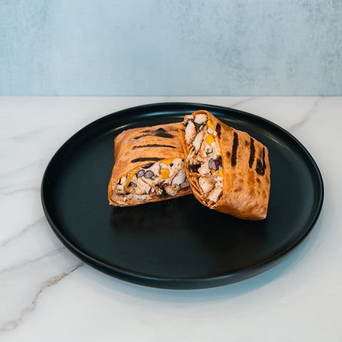 A Broritto Burrito, grilled and cut in half on a black plate, reveals a hearty filling of beans, corn, and tender pieces of chicken. This high-protein burrito rests atop a white marble surface with a light gray background.