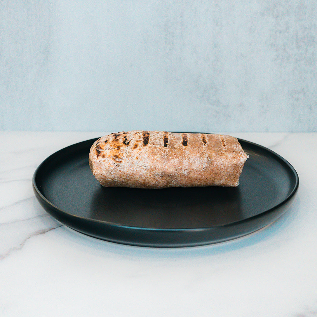 The Arches Mac Daddy Wrap, a high-protein meal prep option inspired by cheeseburger flavors, features grilled char marks and is served on a black plate with a light marble surface and pale blue background—perfect for a macro-friendly lunch.