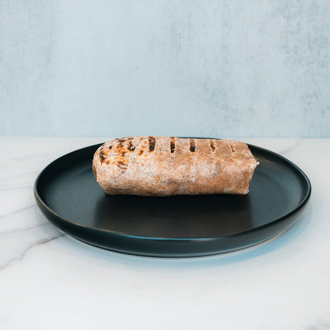 The Arches Mac Daddy Wrap, a high-protein meal prep option inspired by cheeseburger flavors, features grilled char marks and is served on a black plate with a light marble surface and pale blue background—perfect for a macro-friendly lunch.
