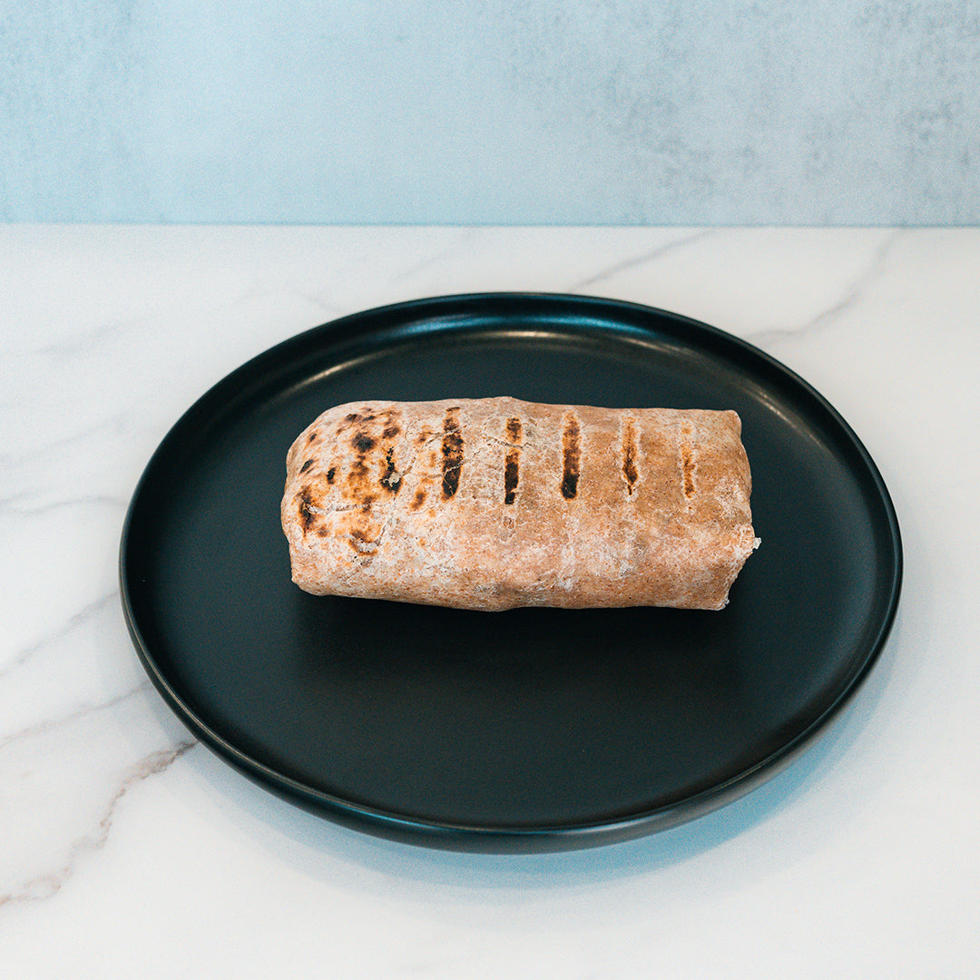 The Arches Mac Daddy Wrap, a grilled wrap with signature grill marks, sits on a round black plate atop a white marble surface—delivering a high-protein meal prep option perfect for a macro-friendly lunch or anyone craving classic cheeseburger-inspired flavor.