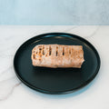 The Arches Mac Daddy Wrap, a grilled wrap with signature grill marks, sits on a round black plate atop a white marble surface—delivering a high-protein meal prep option perfect for a macro-friendly lunch or anyone craving classic cheeseburger-inspired flavor.