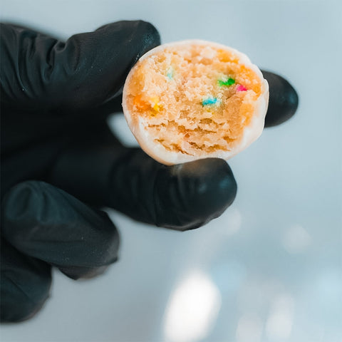 A hand in a black glove holds a bitten Protein Buff Pops - Dunk’d Out, featuring a white coating and colorful sprinkles inside, against a blurred light background—an indulgent yet low calorie protein snack.