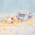 Four white protein cake pops with colorful sprinkles are displayed, one with a bite taken out. Behind them is a clear package labeled Protein Buff Pops - Dunk’d Out. These delicious vanilla cake poppers make the perfect low calorie protein snack, topped with vibrant sprinkles.