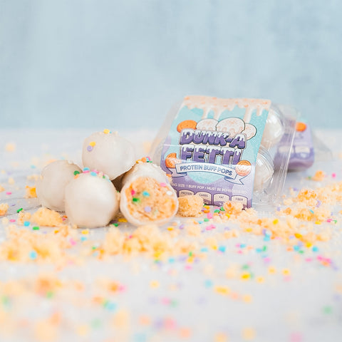 Four white protein cake pops with colorful sprinkles are displayed, one with a bite taken out. Behind them is a clear package labeled Protein Buff Pops - Dunk’d Out. These delicious vanilla cake poppers make the perfect low calorie protein snack, topped with vibrant sprinkles.
