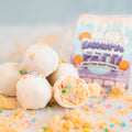 White cake balls with colorful sprinkles are stacked, one with a bite taken out to reveal a soft interior—reminiscent of a classic vanilla cake popper. Crumbs and sprinkles are scattered around, while a blurred box labeled Protein Buff Pops - Dunk’d Out sits in the background, hinting at this fun low calorie protein snack.