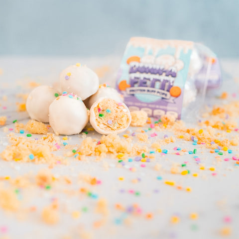 Several white cake balls covered in colorful sprinkles are scattered on a light surface. One Protein Buff Pops - Dunk’d Out cake pop is cut open, revealing a funfetti interior. In the background, a package labeled Protein Buff Pops - Dunk’d Out beckons with the promise of this irresistible low calorie treat.