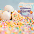 A close-up of Protein Buff Pops - Dunk’d Out, white chocolate-coated vanilla cake popper balls with colorful sprinkles—one sliced open to reveal a soft, sprinkle-filled center. The background features the pastel Protein Buff Pops - Dunk’d Out snack package surrounded by scattered sprinkles, emphasizing this low calorie treat.