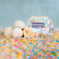 Four white chocolate-covered cake pops with colorful sprinkles are arranged on a surface covered in sprinkles and cake crumbs. Perfect as a low-calorie protein snack, these vanilla cake popper treats come in a clear plastic pack labeled Protein Buff Pops - Dunk’d Out, visible in the background.