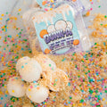 Protein Buff Pops - Dunk’d Out are pictured as white, protein-packed pops coated in sprinkles and crumbs, arranged beside their vibrant festive packaging. Colorful confetti is scattered across a light surface, highlighting these vanilla cake popper-inspired snacks—an irresistibly delicious, low-calorie treat ideal for any celebration.