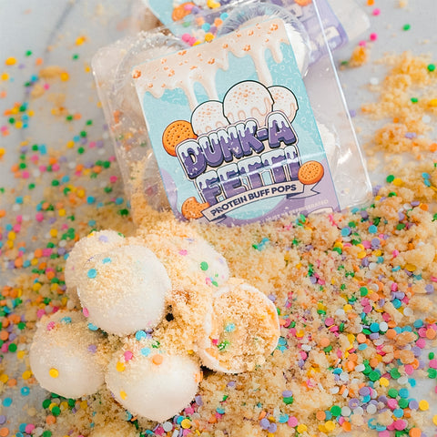 Protein Buff Pops - Dunk’d Out are pictured as white, protein-packed pops coated in sprinkles and crumbs, arranged beside their vibrant festive packaging. Colorful confetti is scattered across a light surface, highlighting these vanilla cake popper-inspired snacks—an irresistibly delicious, low-calorie treat ideal for any celebration.