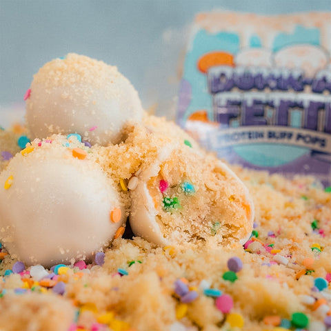 Three round, white Protein Buff Pops - Dunk’d Out cake poppers with colorful sprinkles rest on a bed of cookie crumbs. One popper is cut open to reveal a vibrant funfetti interior, making this protein snack both delightful and playful. In the background, a blurred package of Dunk’d Out low calorie protein snacks can be seen.