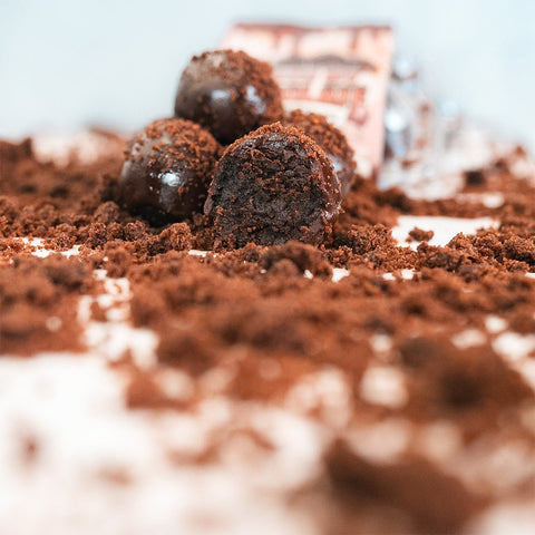 Three Protein Buff Pops—Death By Chocolate, one with a tempting bite taken out—rest on a white surface dusted with chocolate crumbs, making them an ideal grab-and-go treat. A blurred Protein Buff Pops wrapper sits in the background.