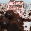 A package of Protein Buff Pops - Death By Chocolate sits among scattered chocolate crumbs, with several round chocolate cake treats in the foreground and plastic packaging visible in the background—perfect for a delicious grab-and-go protein snack.