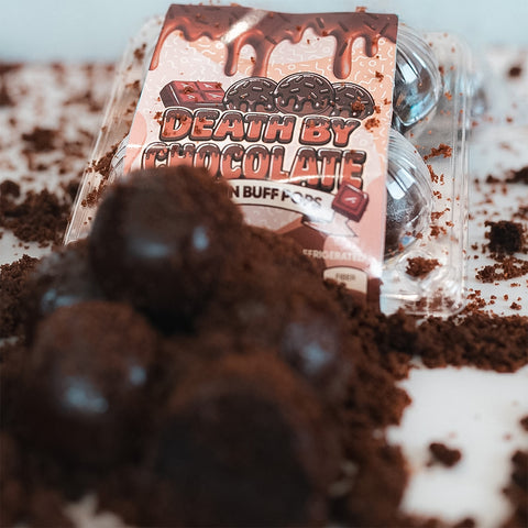 A package of Protein Buff Pops - Death By Chocolate sits among scattered chocolate crumbs, with several round chocolate cake treats in the foreground and plastic packaging visible in the background—perfect for a delicious grab-and-go protein snack.